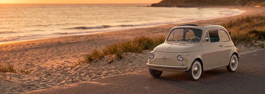 Rivivente Fiat 500 beside the sea at sunset, with a lighthouse in the distance.